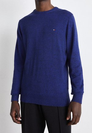 Homme portant un pull à col rond manches longues bleu roi avec un petit logo rectangulaire sur la poitrine et un pantalon noir.