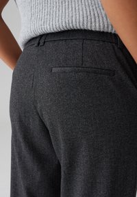 Pantaloni grigi scuri a fantasia pied-de-poule con vestibilità sartoriale. Presentano una tasca posteriore e una consistenza liscia, abbinati a un maglione grigio a coste.