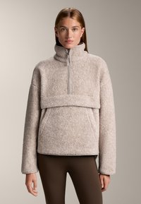 Fleece pullover in lichtbeige met een hoge kraag, ritsluiting en een voorzak. Zachte textuur en oversized mouwen. Bruine leggings.
