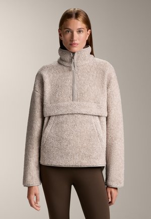 Fleece pullover in lichtbeige met een hoge kraag, ritsluiting en een voorzak. Zachte textuur en oversized mouwen. Bruine leggings.