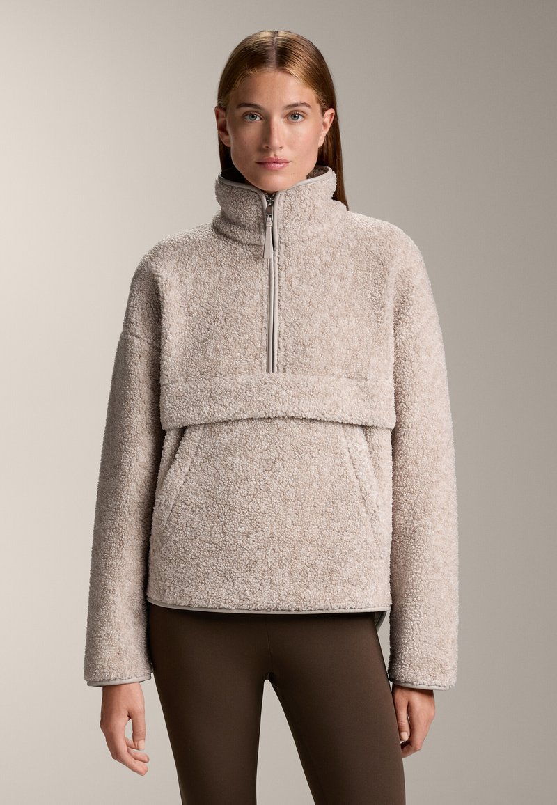 Fleece pullover in lichtbeige met een hoge kraag, ritsluiting en een voorzak. Zachte textuur en oversized mouwen. Bruine leggings.