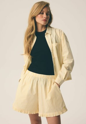 Blondynka ubrana w czarny top, żółto-białą pasiastą koszulę na guziki oraz pasujące luźne szorty z falbankowymi brzegami, ręce w kieszeniach.
