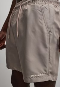 Shorts beiges à cordon avec poches latérales et logo "PEGADOR" brodé près de l'ourlet sur une personne à la peau foncée.