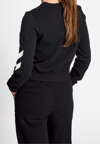 Sudadera negra de manga larga con acentos de chevron blancos en el brazo izquierdo. Cuello y dobladillo acanalados, combinados con pantalones negros a juego.