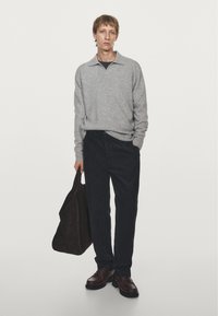Grå stickad tröja med krage, mörka denimbyxor, bruna läderskor och en mörk axelväska, stående mot en enfärgad bakgrund.