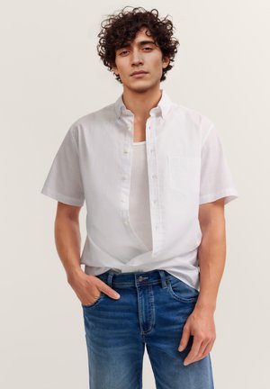 Junger Mann mit lockigem Haar, der ein offenes weißes Kurzarmhemd über einem weißen Tanktop und blauen Jeans trägt, steht mit einer Hand in der Hosentasche.