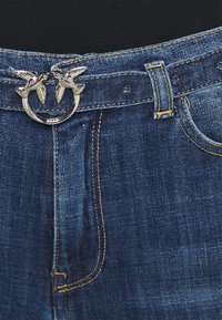 Jeans de denim con un lavado azul oscuro. Presenta una hebilla de cinturón plateada en forma de dos pájaros y costuras amarillas a lo largo de las costuras.