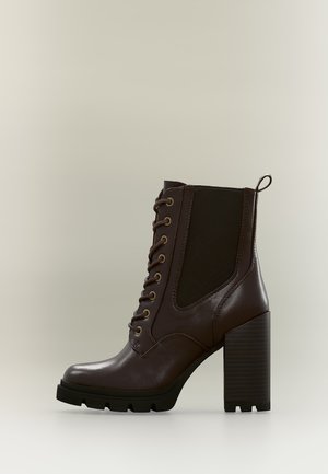 Bottine en cuir marron foncé avec fermeture à lacets sur l’avant, talon haut épais et empiècement latéral élastique noir.