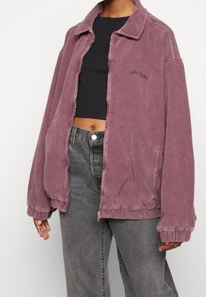 Person wearing oversized dusty purple corduroy zip-up jacket over black cropped top and faded black jeans, standing against white background.