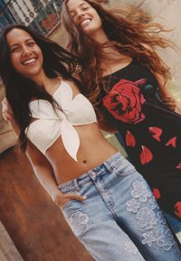 Két nő áll egymás mellett. Az egyik egy fehér, virágmintás cropped topot és világoskék, virágos hímzésű farmert visel. A másik egy fekete ruhát visel, piros rózsa mintával.