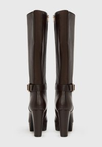 Σκούρες καφέ δερμάτινες μπότες έως το γόνατο με φαρδιές φτέρνες, διακοσμητικές πλευρικές λουρίδες και αγκράφες σε χρυσό τόνο, εμφανιζόμενες από πίσω πάνω σε απλό φόντο.