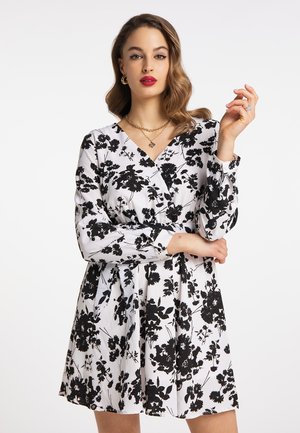 Jeune femme aux cheveux ondulés porte une robe blanche avec un imprimé floral noir, un rouge à lèvres rouge, des boucles d'oreilles dorées et des colliers superposés, posant les bras croisés.