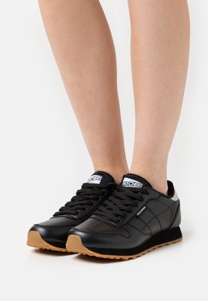 Voeten met zwarte Skechers vetersneakers met gumzolen tegen een effen witte achtergrond.