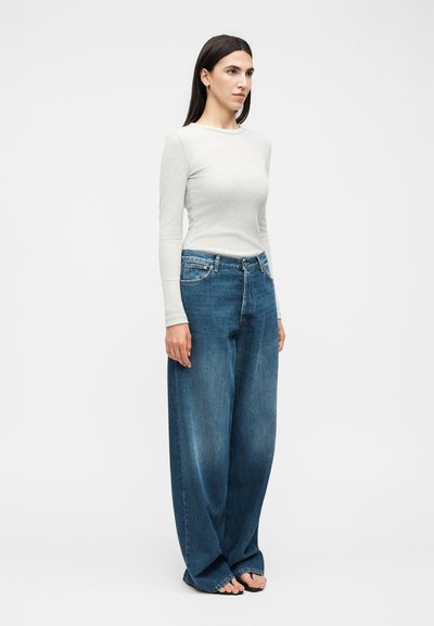 Γυναίκα στέκεται, φοράει εφαρμοστή λευκή μπλούζα με μακριά μανίκια και φαρδιά μπλε τζιν με σανδάλια φλίπ-φλοπ, σε απλό φόντο.