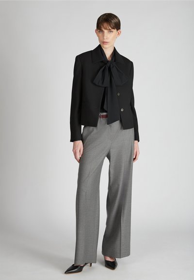 Giacca nera cropped con un grande fiocco, pantaloni grigi a gamba larga e décolleté nere a punta; tessuto elegante e design strutturato.