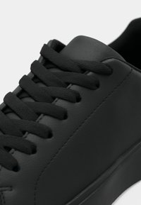 Fekete, alacsony szárú sportcipő fekete fűzővel, sima bőr textúrával és gumitalppal, fehér háttéren.