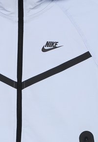 Ljusblå jacka med svarta accenter, utrustad med dragkedja och en framträdande Nike-logotyp. Slät textur och modern design med geometriska linjer.