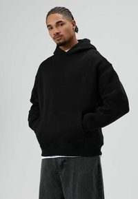 Schwarzer Kapuzenpullover mit Fronttasche, gerippten Bündchen und einem dezenten Logodetail. Über einem weißen Hemd getragen, kombiniert mit dunklen Jeans.