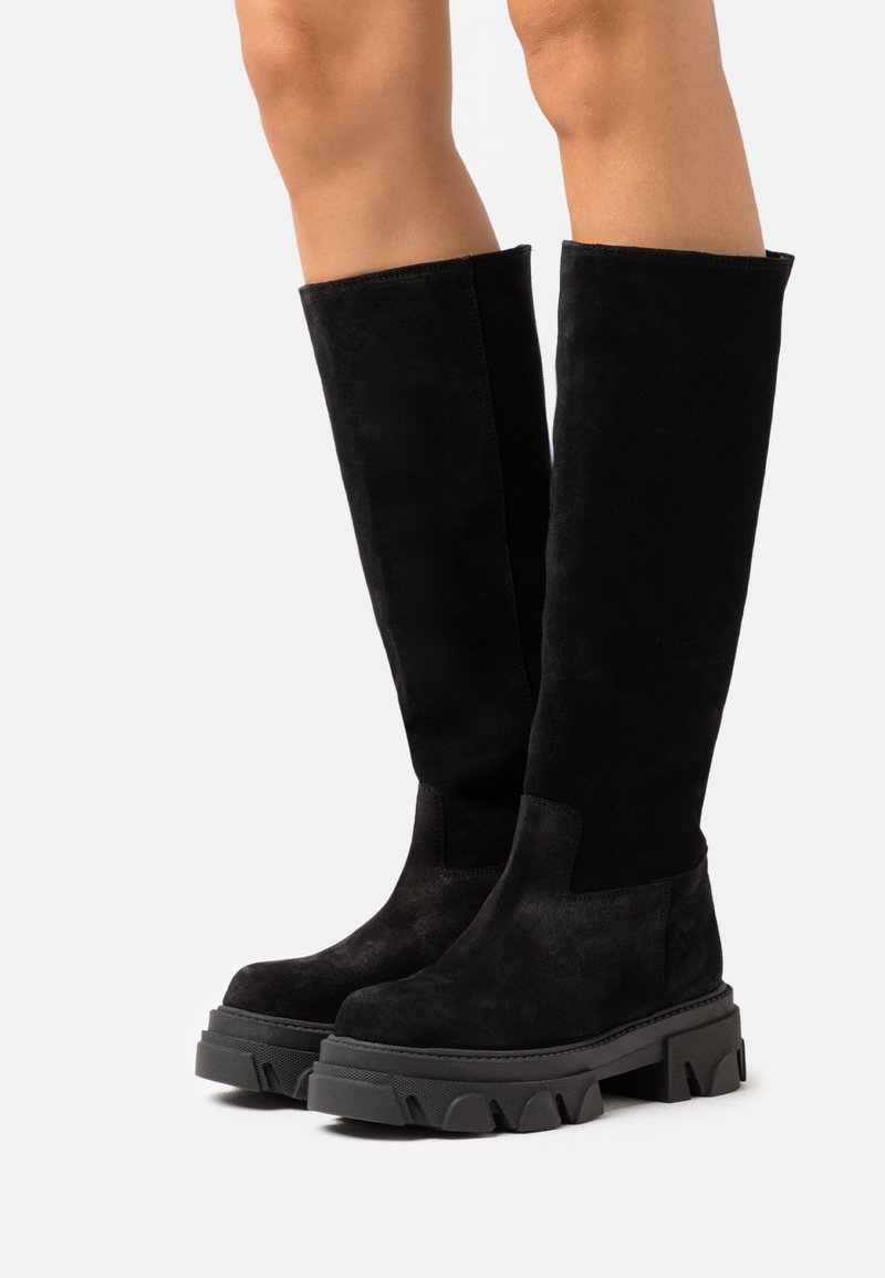 Bottes hautes jusqu'au genou en daim noir avec semelle en caoutchouc noire épaisse et rainures prononcées. Présentent une texture lisse et des panneaux latéraux élastiques pour un ajustement optimal.