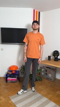 Orange t-shirt med en liten logotyp, grå jeans och vita sneakers. Bakgrunden har en TV, en lampa och färgglada väggstripes.