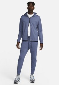 Trainingspak in lichtblauwe stof met een hoodie met ritssluiting, bijpassende broek met trekkoord en zwarte accenten. Draagt een paar grijze en zwarte sneakers.