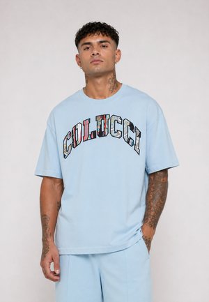 Mężczyzna z tatuażami, ubrany w jasnoniebieski t-shirt Colucci i pasujące spodnie, stoi na jednolitym tle.