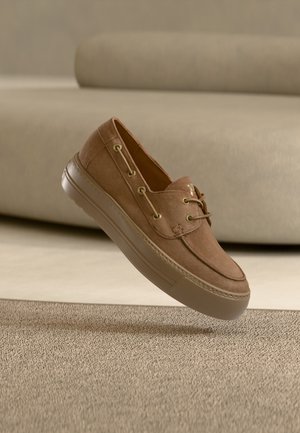 Chaussure décontractée en daim marron avec des lacets flottant au-dessus d'un tapis beige, avec un canapé marron clair en arrière-plan.