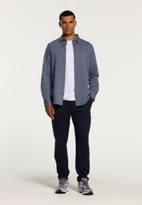 Svetlo modra srajca z dolgimi rokavi in gumbi čez belo majico, v kombinaciji s temno mornarskimi trenirkami in sivimi športnimi čevlji. Enostaven dizajn.