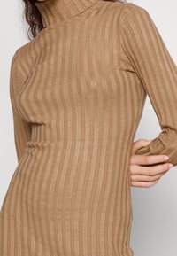 Maglia a maniche lunghe con collo alto in tessuto a coste color sabbia, caratterizzata da strisce verticali e una silhouette aderente. Texture morbida e materiale elasticizzato.