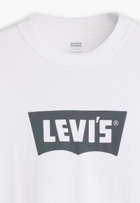 Balta medvilninė marškinė su pilku Levi's logotipu išskirtine kreiva forma. Klasikinis apvalus iškirptė, minkšta tekstūra, senovinė forma.