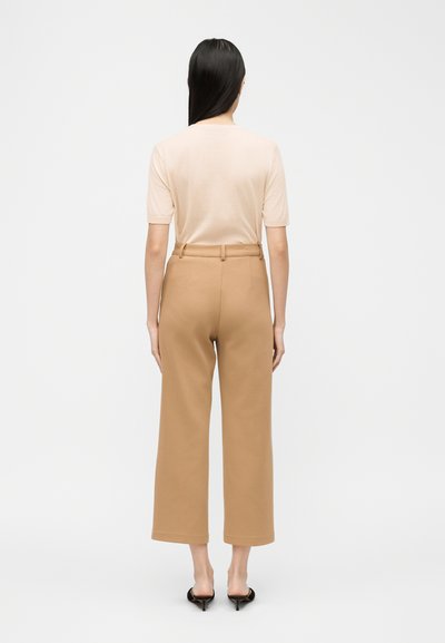 Γυναίκα με μακριά σκούρα μαλλιά φοράει μπεζ τοπ με κοντά μανίκια, παντελόνι καμηλό με ψηλή μέση και φαρδιά πατζάκια, και μαύρα mule με τακούνι, εμφανιζόμενη από πίσω.