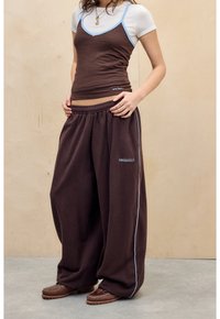 Bruna, löst sittande sweatpants med elastiskt midjeband och blåa detaljer, i kombination med en brun linne med blå accenter och en vit skjorta under.