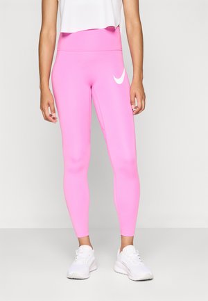 Woman wearing bright pink Nike leggings and white sneakers, standing against a plain light gray background.