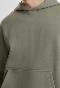 Felpa con cappuccio verde oliva in tessuto morbido, con tasca frontale stile canguro e cappuccio con coulisse. Tessuto liscio e vestibilità comoda.