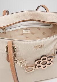 Beżowa torba skórzana z teksturowanej skóry, zapinana na zamek, z kremową podszewką oraz logo 'GUESS'. Posiada złotą łańcuszek z kwiatowym zawieszką.
