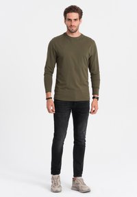 Olivegrünes Langarmshirt mit Rundhalsausschnitt, kombiniert mit dunklen Jeans und beigen Sportschuhen. Schlichtes, tailliertes Design.