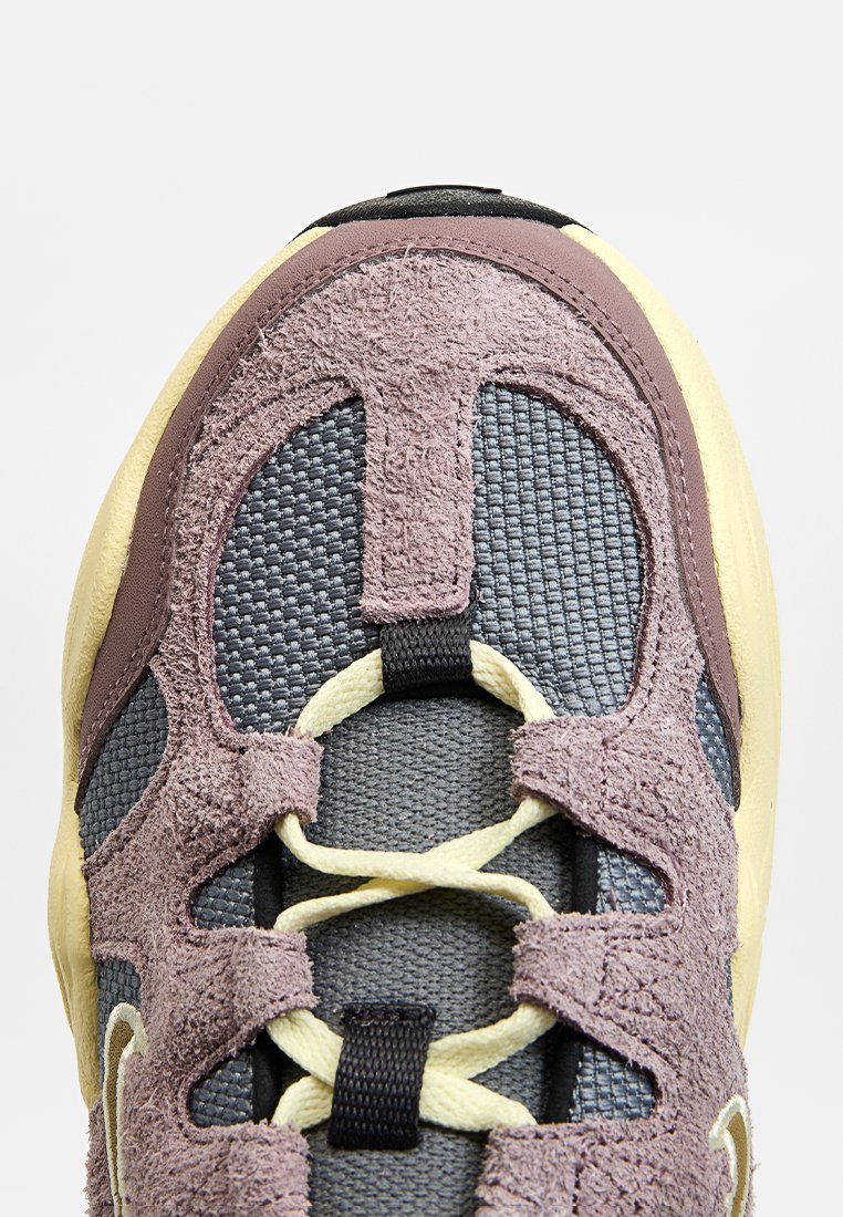 Sneaker z połączeniem szarej siateczki i fioletowego zamszu. Posiada kontrastujące żółte sznurowadła i podeszwę, z teksturowanym, nowoczesnym wzornictwem.