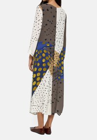 Kobieta w długiej, luźnej sukience z abstrakcyjnymi wzorami w niebieskie, złote, białe i brązowe kropki, zestawione z brązowymi płaskimi butami.