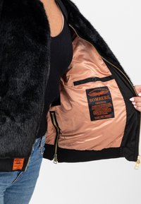 Giacca bomber in finta pelliccia nera con un interno lucido, zip dorata e una tasca interna con un'etichetta arancione contenente istruzioni per la cura.