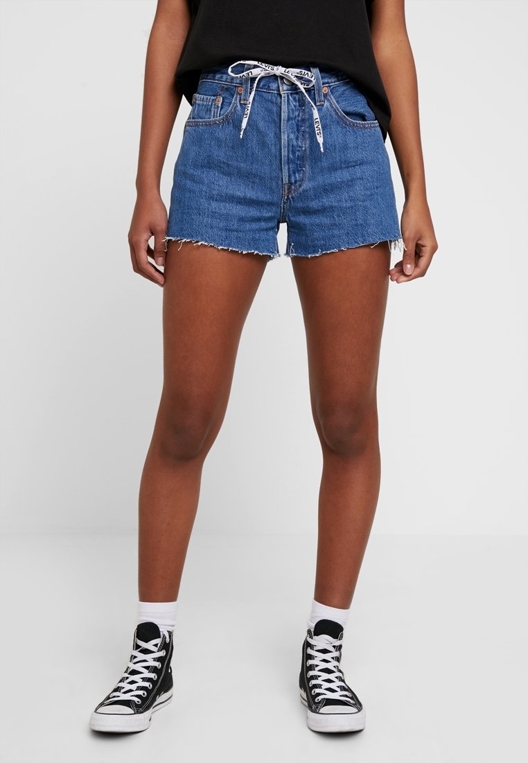 Person wearing blue denim shorts with frayed hem and white Levi's drawstring, black high-top sneakers, white socks, standing against plain background.