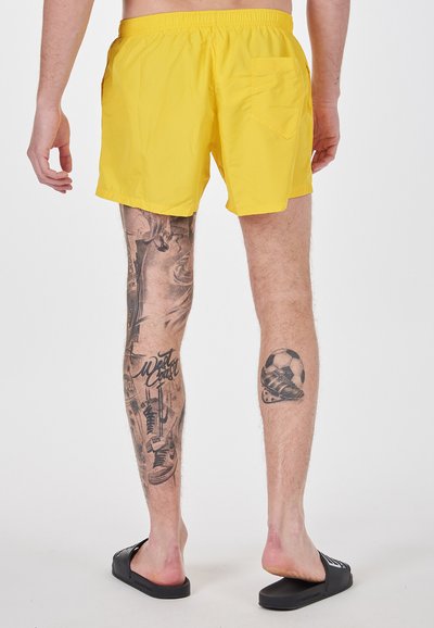 Costumi da bagno gialli con cintura elastica, tasche laterali e tasca sul retro. Presenta tatuaggi visibili sulle gambe e sandali neri.