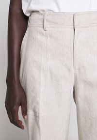 Ljusbeige linbyxor med rak bendesign, med sidofickor och en slät yta. Bältesömmar är synliga i midjan.