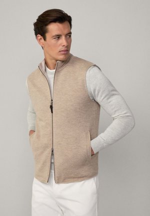 Beige ärmellose Weste aus weichem Strickmaterial, mit einem hohen Kragen, einem Reißverschluss vorne und seitlichen Taschen, über einem hellgrauen Langarmshirt getragen.