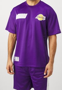 Mężczyzna w fioletowej koszulce koszykarskiej Los Angeles Lakers NBA i dopasowanych spodenkach z białymi logo na jednolitym tle.
