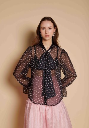 Vrouw met steil bruin haar draagt een doorschijnende zwarte blouse met polka dots en een das over een zwarte top en een gestructureerde roze rok, staand tegen een beige muur.
