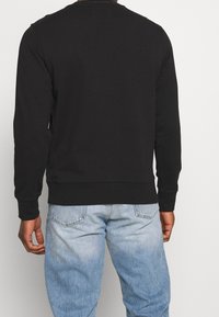 Sweatshirt noir à manches longues, avec poignets et ourlet côtelés. Présente un logo au niveau du col. Porté avec un jean en denim clair et délavé.