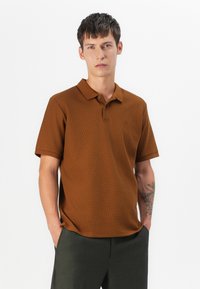 Smeđi teksturirani polo majica s klasičnom ovratnikom, kratkim rukavima i suptilnim logotipom, u kombinaciji s maslinasto-zelenim hlačama.