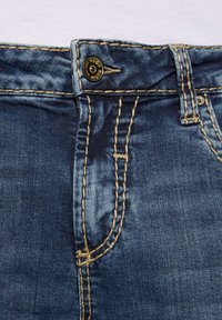 Jeans aus Denim mit einer dunkelblauen Waschung, die mit goldenen Nähten und einem Knopfverschluss am Bund versehen sind, zeigen eine glatte Textur und detaillierte Nähte.
