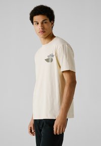 Camiseta de algodón color crema con mangas cortas, que presenta un gráfico impreso en el lado izquierdo del pecho y un corte relajado. Combinada con jeans oscuros.