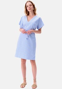 Wzorzysty niebiesko-biały sukienka z dekoltem w kształcie litery V i krótkimi, falbaniaste rękawami, podkreśloną talią i długością do kolan. Założona z złotymi sandałami na cienkich paskach.